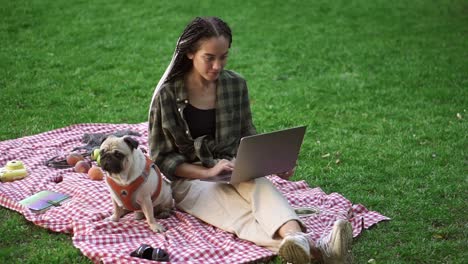 Stylish-Girl-Sitting-Outdoors-On-The-Grass-Lawn-With-Her-Laptop-Computer-And-Cute-Little-Pug-Is-Sitting-Near