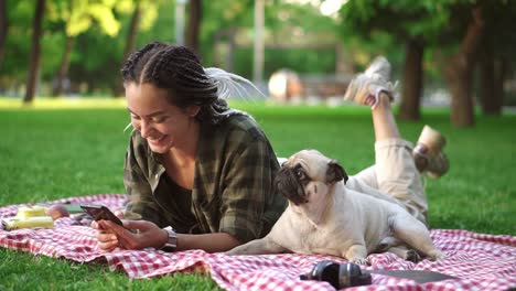 Woman-Laying-Outdoors-On-The-Picnic-Plaid-On-Lawn-Using-Her-Smartphone-And-Cute-Little-Pug-Is-Laying-Near