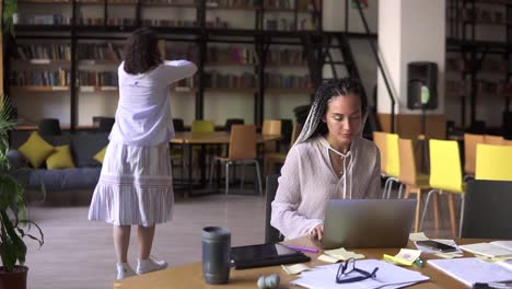 Happy-Brunette-In-White-Casual-Clothes-In-Headphones-Funny-Dancing-On-Background-While-Her-Friend-Concentrated-On-Her-Studying,-Sitting-At-The-Table-Works-With-Laptop