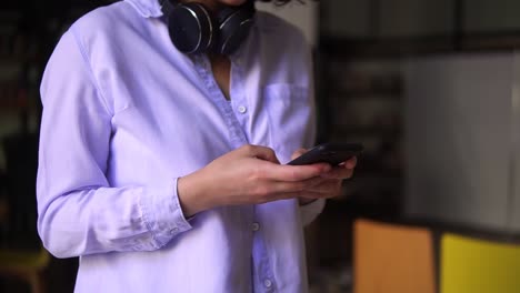 Cropped-Footage-Of-Walking-Brunette-Woman-Wearing-White-Blouse,-Looking-On-Smartphone-Screen-In-Her-Hands-And-Typing,-Headphones-On-Neck