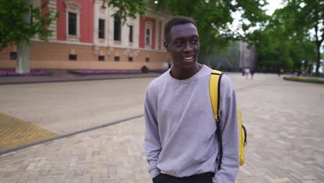 Positive,-Smiling-Young-Man-Walking-In-City-With-Yellow-Backpack-On-Shoulders,-Wearing-Casual-Clothes