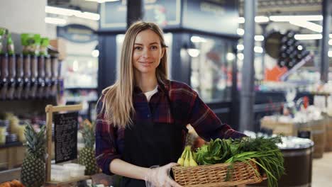 Female-Supermarket-Employee-In-Black-Apron-Holding-A-Box-Full-Of-Fruits-And-Greens-In-Supermarket
