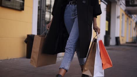 Lose-Up-Of-Woman-Legs-Wearing-Stylish-Black-Coat-And-Jeans-At-Day-Time-Hold-Colored-Shopping-Bags-After-Big-Shopping-Day