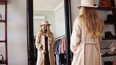 Attractive-Blonde-Girl-Standing-In-Front-The-Mirror-In-Beige-Coat-And-Trendy-Hat,-Making-A-Photo-Of-Look-With-Her-Smartphone