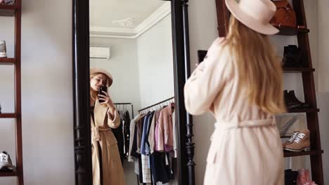 Fashionable-Blonde-Girl-Standing-In-Front-The-Mirror-In-Beige-Coat-And-Trendy-Hat,-Making-A-Photo-Of-Look-With-Her-Smartphone