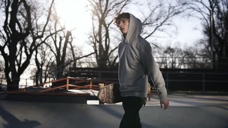 Young-Skateboarder-In-Grey-Hoodie-Walks-In-A-Skate-Park-With-A-Skateboard-In-Hands