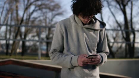 Young-Curly-Headed-Male-Skateboarder-Browsing-In-His-Mobile-Phone-In-The-Skate-Park-On-A-Sunny-Day-1