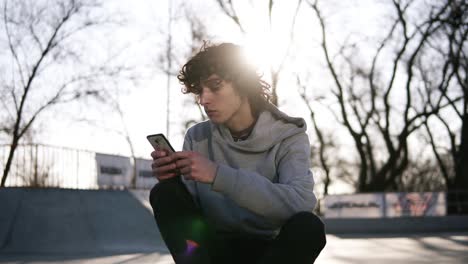 Young-Man-Is-Holding-A-Mobile-Phone-While-Sitting-In-A-Skate-Zone-In-Skate-Park