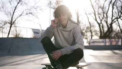 Young-Man-Is-Calling-To-Friends-Using-Mobile-Phone-While-Sitting-In-A-Skate-Zone-In-Skate-Park