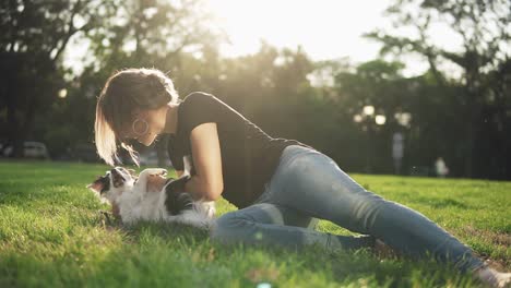 Front-Footage-Of-A-Woman-In-Jeans-And-Black-T-Shirt-Lying-On-The-Green-Grass-In-The-Park-And-Playing-With-Her-Pet