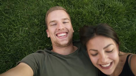 Young-In-Love-Heterosexual-Couple-Resting-Outdoors-In-Park-On-Lawn,-Green-Grass-Lying-In-Embrace-And-Making-Selfie-Photo-On-Smartphone-Camera
