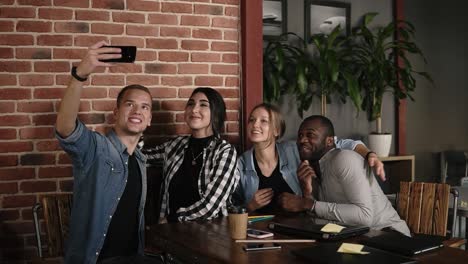 Man-Taking-Group-Selfie-At-Meeting-With-Diverse-Friends-In-Coffeeshop-Or-Workplace,--Positive-Young-People-Making-Photo-Together-In-Cafe-With-Brick-Wall-Interior,-Multi-Ethnic-Happy-Buisness-People-Photographing-On-Smartphone