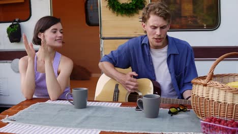 Positive-Couple-Sitting-At-The-Picnic-Table-Outdoors-In-Front-Their-Home-Trailer-And-Man-Playing-The-Guitar-And-Sings-Song-Actively