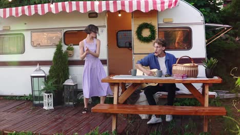 Funny-Girl-In-Blue-Summer-Dress-Girl-Dancing-While-Man-Playing-Guitar-Sitting-At-The-Wooden-Table-In-Front-Trailer,-Singing-A-Song-Loudly