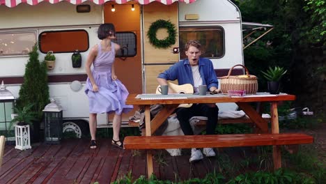 Brunette-Girl-In-Blue-Summer-Dress-Girl-Dancing-While-Man-Playing-Guitar-Sitting-At-The-Wooden-Table-In-Front-Trailer,-Singing-A-Song-Loudly