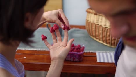 Young-Woman-Sitting-Together-With-Her-Boyfriend-At-The-Wooden-Table,-Putting-Raspberries-On-Her-Fingers-Smiling-And-Posing