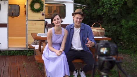 Popular-Bloggers-Couple-Recording-Video-About-Themseves,-Sitting-At-The-Wooden-Table-In-Front-The-Wheels-House,-Talking-And-Looking-At-Camera-On-Tripod