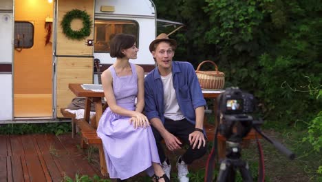 Popular-Bloggers-Couple-Greeting-Their-Followers,-Recording-Video-While-Sitting-At-The-Wooden-Table-In-Front-The-Wheels-House,-Talking-And-Looking-At-Camera-On-Tripod