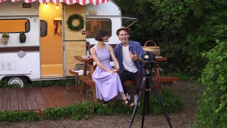 Popular-Bloggers-Couple-Greeting-Their-Followers,-Recording-Video-While-Sitting-At-The-Wooden-Table-In-Front-The-Wheels-House,-Talking-And-Looking-At-Camera-On-Tripod-1
