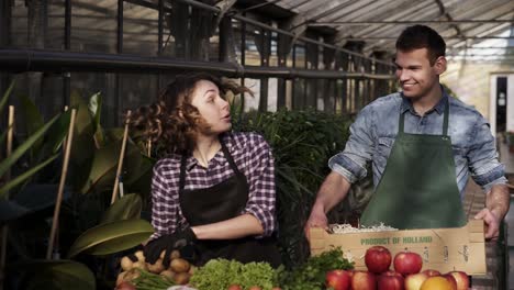 Slow-Motion-Footage-Of-A-Handsome,-Tall-Man-In-Green-Apron-Carrying-The-Box-With-Fresh-Unrecognizable-Vegetables-And-Fruits-To-The-Woman-Selling-Organic-Products