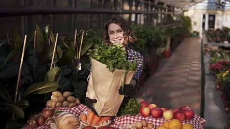 Young-Woman-In-Apron-Holding-Brown-Paper-Bag-With-Fresh-Vegetables-And-Greens,-Smiling-Laughing-While-Posing-For-Camera-In-Food-Market-Standing-In-A-Row-Of-Indoors-Greenhouse-In-Front-The-Table-With-Organic-Food