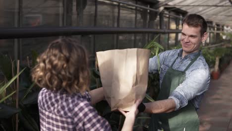 Positive,Tall-Salesman-In-Green-Apron-Giving-Fresh-Vegetables-In-Paper-Bag-To-Customer-In-Greenhouse-Market