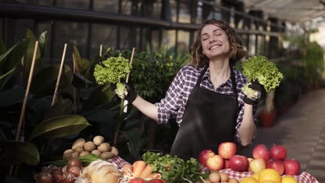 Overjoyed-Young-Seller-In-Black-Apron-Standing-In-A-Row-Of-Greenhouse-In-Front-The-Table-With-Groceries,-Holding-In-Hands-Two-Piles-Of-Green,-Fresh-Salad-And-Making-Funny-Dance,-Smiling-And-Excited