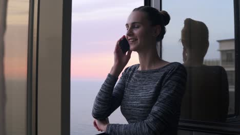Young-Smiling-Woman-Dialing-A-Number-And-Starting-To-Speak-On-The-Phone-While-Standing-By-The-Open-Window-With-A-Smile-During-The-Sunset-By-The-Sea