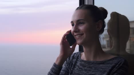 Closeup-View-Of-Young-Smiling-Woman-Speaking-On-The-Phone-While-Standing-By-The-Open-Window-With-A-Smile-During-The-Sunset-By-The-Sea