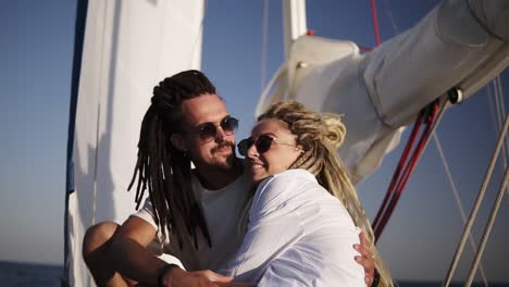 Low-Angle-Footage-Of-Stylish-Couple-With-Dreadlocks-In-White-Clothes-And-Sunglasses-Sitting-Embracing-On-The-Bow-Of-The-Yacht-And-Kissing