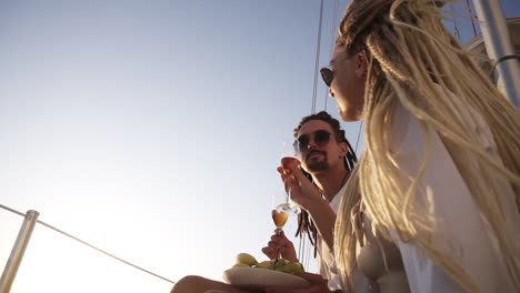 Low-Angle-Footage-Of-Stylish-Couple-With-Dreadlocks-In-White-Clothes-And-Sunglasses-Sitting-Sitting-On-The-Edge-Of-The-Yacht-Smiling,-Clinking-With-Champangne-Glasses