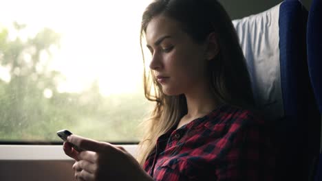 Attractive-Female-In-Plaid-Shirt-Is-Going-By-Train,-Sitting-Next-To-The-Window-And-Seriously-Looking-On-Her-Mobile-Phone