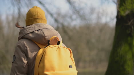 A-Young-Girl-In-A-Yellow-Wool-Cap-And-A-Yellow-Backpack-Walks-Through-The-Forest-Towards-The-Lake