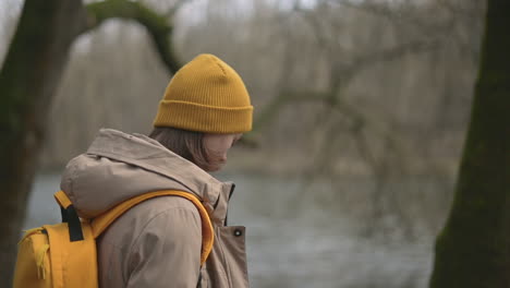 A-Young-Girl-In-A-Forest-That-Wears-A-Yellow-Wool-Cap-And-A-Yellow-Backpack-Walks-Towards-The-Lake