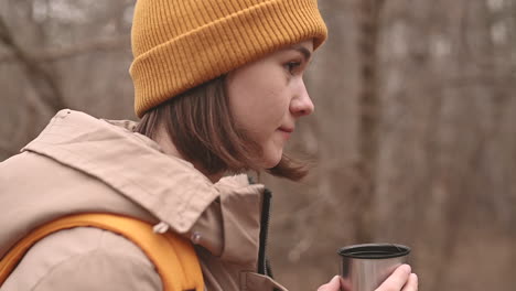A-Young-Girl-In-A-Yellow-Woolen-Hat-Drinks-A-Hot-Beverage-In-The-Forest