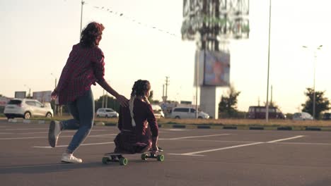 Back-View-Of-A-Woman-Sitting-On-A-Longboard-While-Her-Friend-Is-Pushing-Her-Behind-And-Running-During-Sunset
