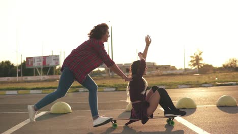 Side-View-Of-A-Woman-With-Her-Hands-Raised-Up-Sitting-On-A-Longboard-While-Her-Friend-Is-Pushing-Her-Behind-And-Running-During-Sunset
