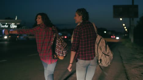 Two-Young-Women-Hitchhikers-Stand-On-The-Sidelines-Holding-Their-Backpacks-And-Ask-To-Stop-The-Passing-Cars-In-The-Night-In-Summertime
