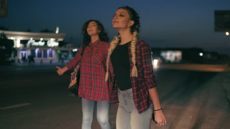 Two-Young-Women-Hitchhikers-Stand-On-The-Sidelines-Holding-Their-Backpacks-And-Ask-To-Stop-The-Passing-Cars-In-The-Night-In-Summertime-1
