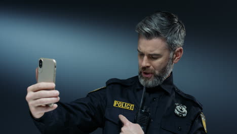 Young-Joyful-Handsome-Policeman-In-Uniform-And-With-Walkie-Talkie-Smiling-And-Having-Videochat-On-The-Smartphone-Via-Web-Cam-On-The-Dark-Wall-Background