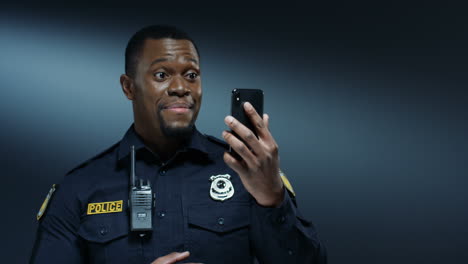 Junger,-Fröhlicher,-Gutaussehender-Polizist-In-Uniform-Und-Im-Walkie-talkie,-Der-Lächelt-Und-Videochat-Auf-Dem-Smartphone-über-Web-cam-Auf-Dem-Dunklen-Wandhintergrund-Hat