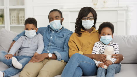 Bored-Young-Family-With-Small-Kids-In-Medical-Masks-Sitting-On-Couch-In-Hugs-And-Looking-At-Camera