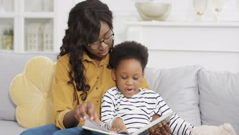 Young-Beautiful-Woman-In-Glasses-Reading-Book-With-Little-Cute-Daughter-On-Sofa-At-Home