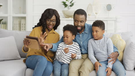 Junge-Familie-Mit-Zwei-Kindern,-Die-Auf-Der-Couch-Im-Wohnzimmer-Sitzen-Und-Auf-Dem-Tablet-gerät-Videochatten