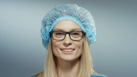 Close-Up-Of-The-Pretty-Female-Young-Doctor-In-Glasses,-Blue-Hat-Smiling-Cheerfully-To-The-Camera