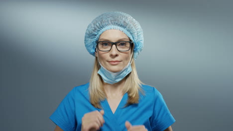 Retrato-De-La-Joven-Y-Atractiva-Doctora-Rubia-Con-Sombrero-Azul-Y-Gafas-Quitándose-La-Máscara-De-La-Boca-Y-Sonriendo-Alegremente