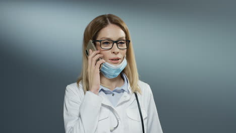 Schöne-Junge-Ärztin-Mit-Brille,-Die-Die-Maske-Vom-Mund-Abnimmt-Und-Fröhlich-Am-Telefon-Spricht-Und-Lächelt