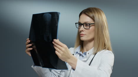 Young-Attractive-Woman-Doctor-Holding-X-Ray-Copy-And-Studying-It-Carefully-On-The-Grey-Background