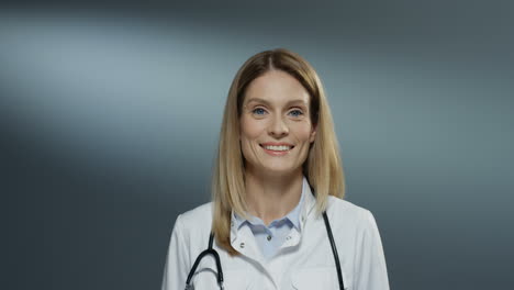 Portrait-Shot-Of-The-Young-Blonde-Pretty-Woman-Physician-In-The-White-Medical-Gown-Turning-To-The-Camera-And-Smiling-Happily-On-The-Gray-Wall-Background