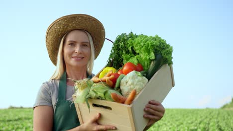Portrait-Of-The-Blonde-Young-Pretty-Woman-Farmer-In-A-Hat-Demonstrating-A-Box-Full-Of-The-Mature-Vegetables-In-The-Green-Field